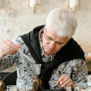 Elderly man carefully crafting a perfume blend in an indoor workspace with various bottles.