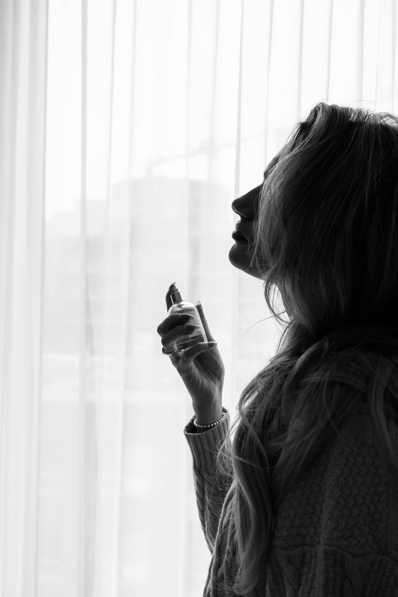 Silhouette of a woman holding and spraying perfume by a window curtain.